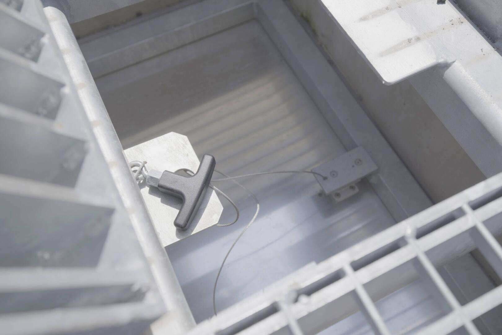 Nahaufnahme eines Metallgitters und von Behältern mit einem schwarzen T-förmigen Werkzeug, das an einem dünnen Draht befestigt ist und in einem rechteckigen Metallfach ruht. Die Szene ist gut beleuchtet, mit Schatten und Spiegelungen.