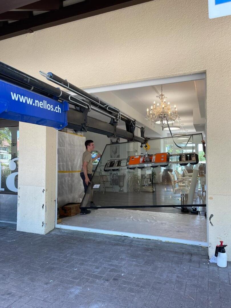 Ein Arbeiter beaufsichtigt eine große Glasscheibe, die mit einem mechanischen Aufzug durch den Eingang eines Gebäudes installiert wird, in dem ein Kronleuchter und moderne Möbel zu sehen sind.
