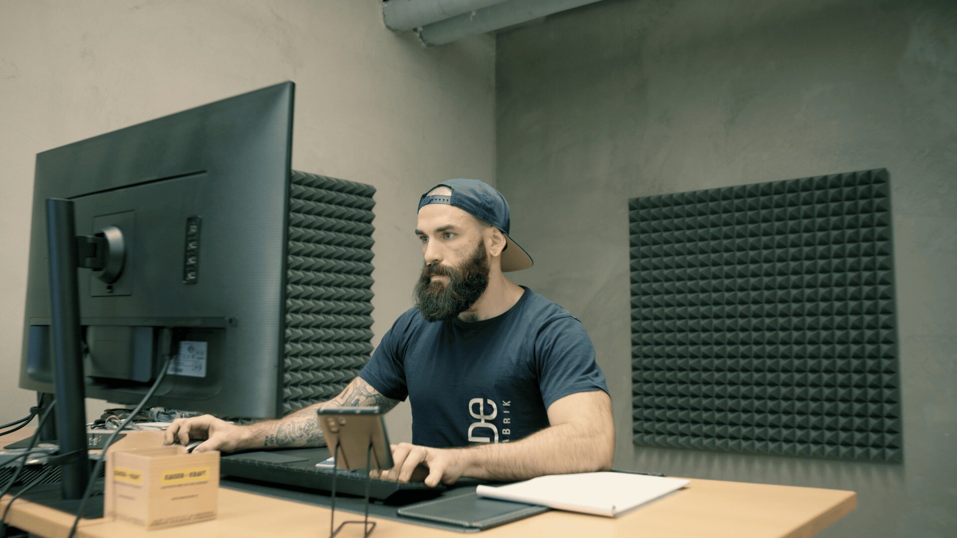Ein bärtiger Mann mit einer Schirmmütze arbeitet an einem Desktop-Computer in einem modernen Büro mit schalldichten Schaumstoffplatten an der Wand. Auf dem Schreibtisch liegen ein Notebook, ein Smartphone und eine Box.