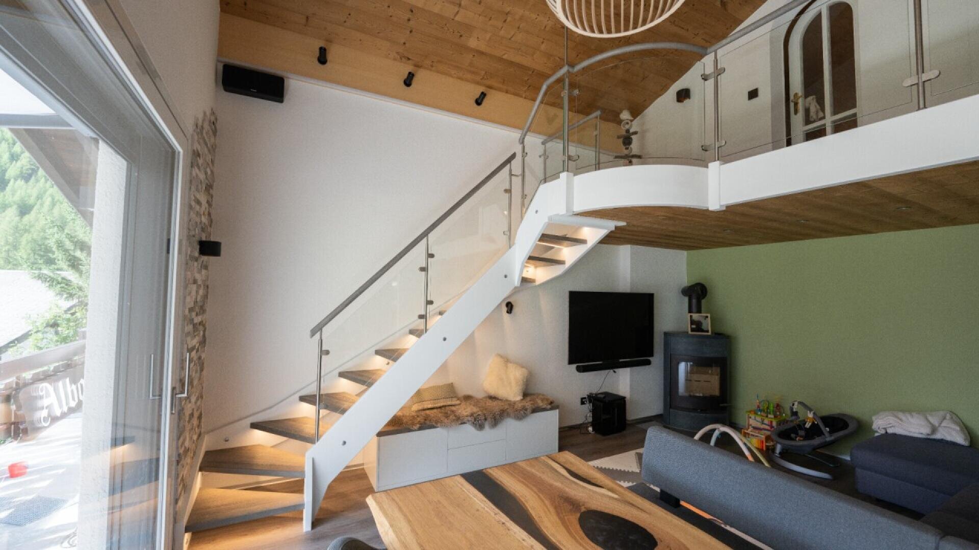 Salon moderne avec un plafond en bois, un loft ouvert, une rampe d'escalier en verre, un mur d'accent vert, un poêle à bois, une télévision et une grande fenêtre qui laisse entrer la lumière naturelle. Une table en bois et des meubles assortis complètent l'espace.
