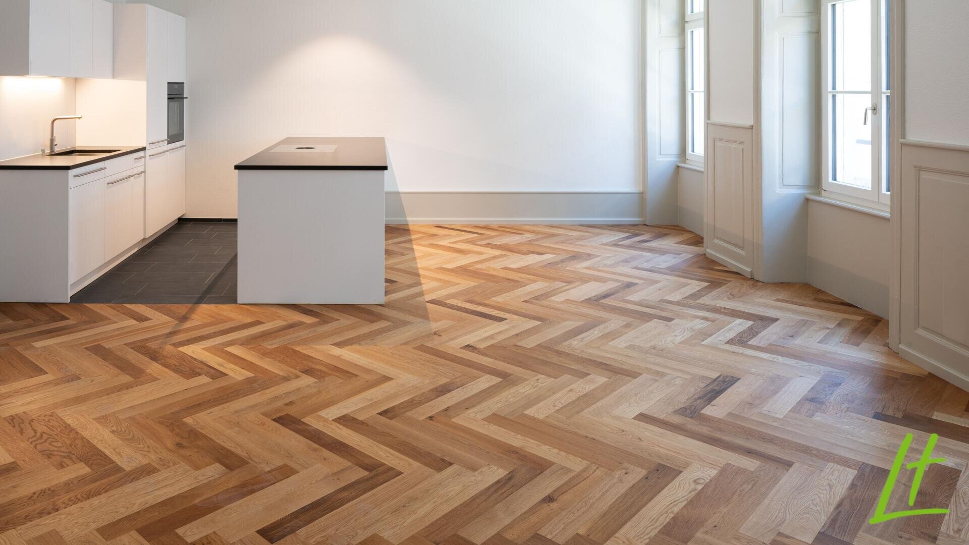 Cuisine et salle à manger modernes et vides avec des armoires blanches, des plans de travail noirs et un îlot central. La pièce dispose de grandes fenêtres et de parquets en bois clair avec des motifs à chevrons.