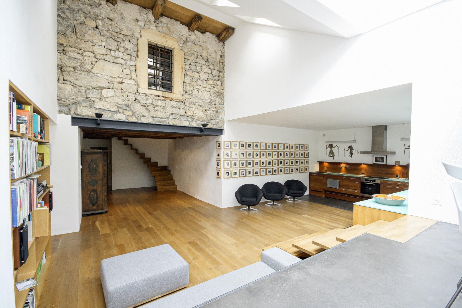 Moderner offener Wohnraum mit Holzböden, freiliegender Steinwand, eingebautem Bücherregal, vier schwarzen Stühlen, einem Küchenbereich und einer Treppe, die zu einem oberen Stockwerk mit einem rustikalen Fenster führt.