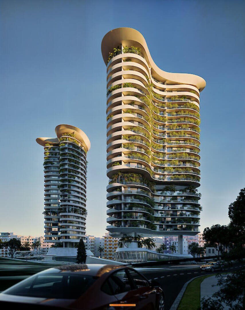 Deux immeubles modernes aux lignes courbes, aux façades de verre et à la végétation luxuriante sur les balcons, se côtoient au crépuscule, dans un ciel dégagé. Sur la route au premier plan, une voiture passe.