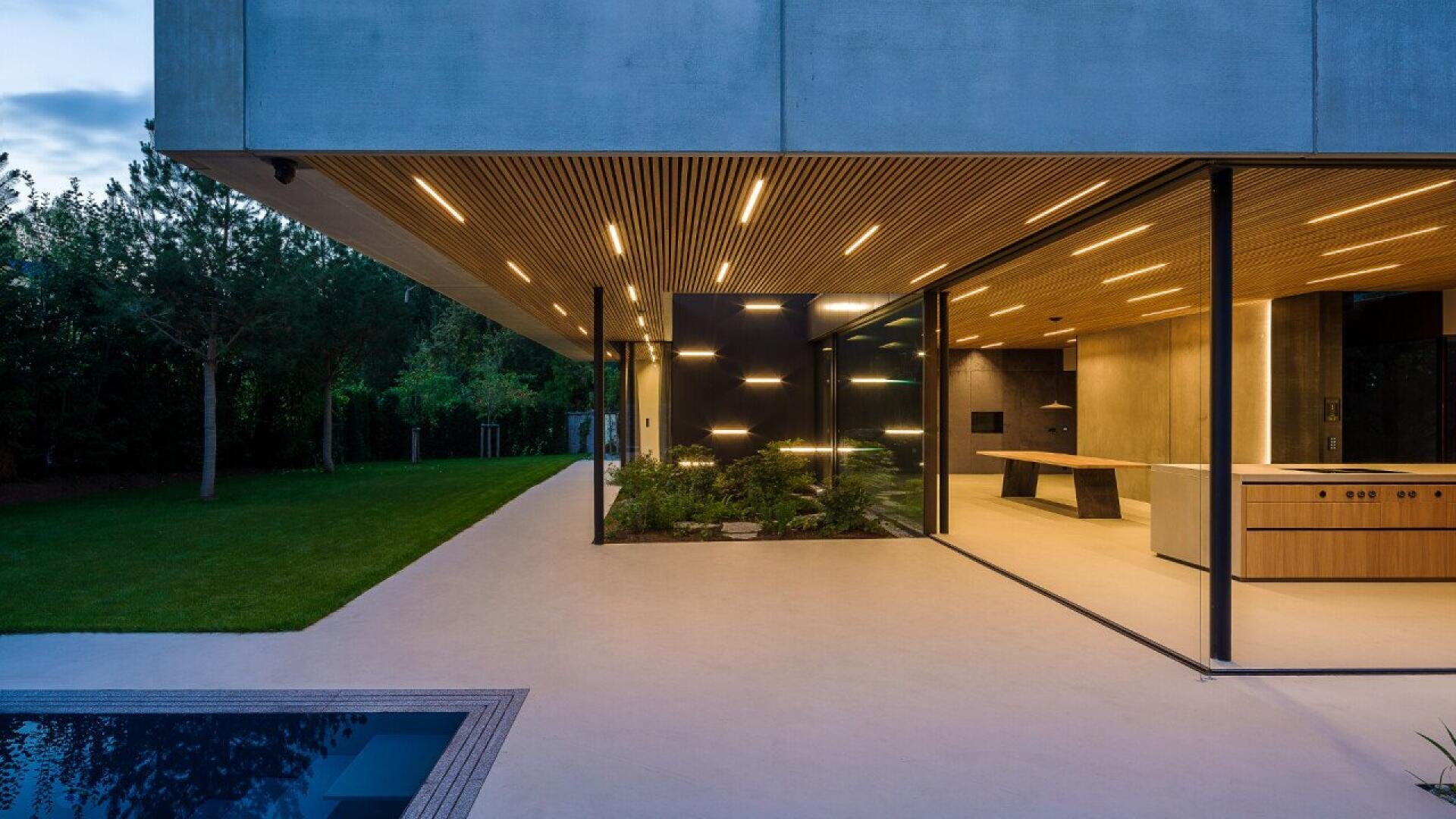 Maison moderne en béton avec un plafond en lattes de bois et des luminaires intégrés, donnant sur une pelouse et une piscine. Les murs en verre révèlent une cuisine et une salle à manger contemporaines.