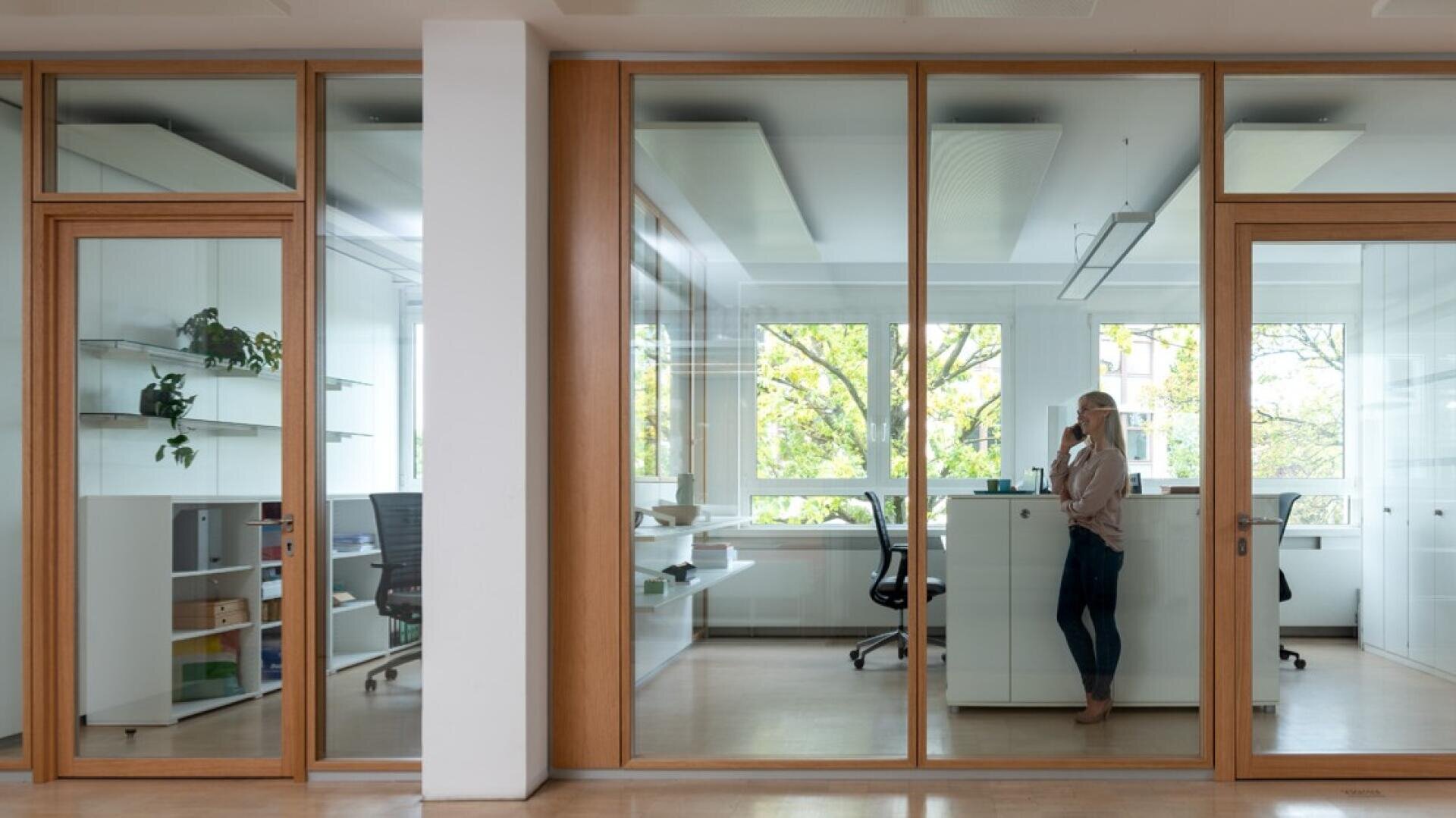 Une personne se tient dans un bureau moderne et lumineux avec des murs en verre et parle au téléphone. Il y a des bureaux, des chaises de bureau, des étagères et de grandes fenêtres qui donnent sur la verdure.