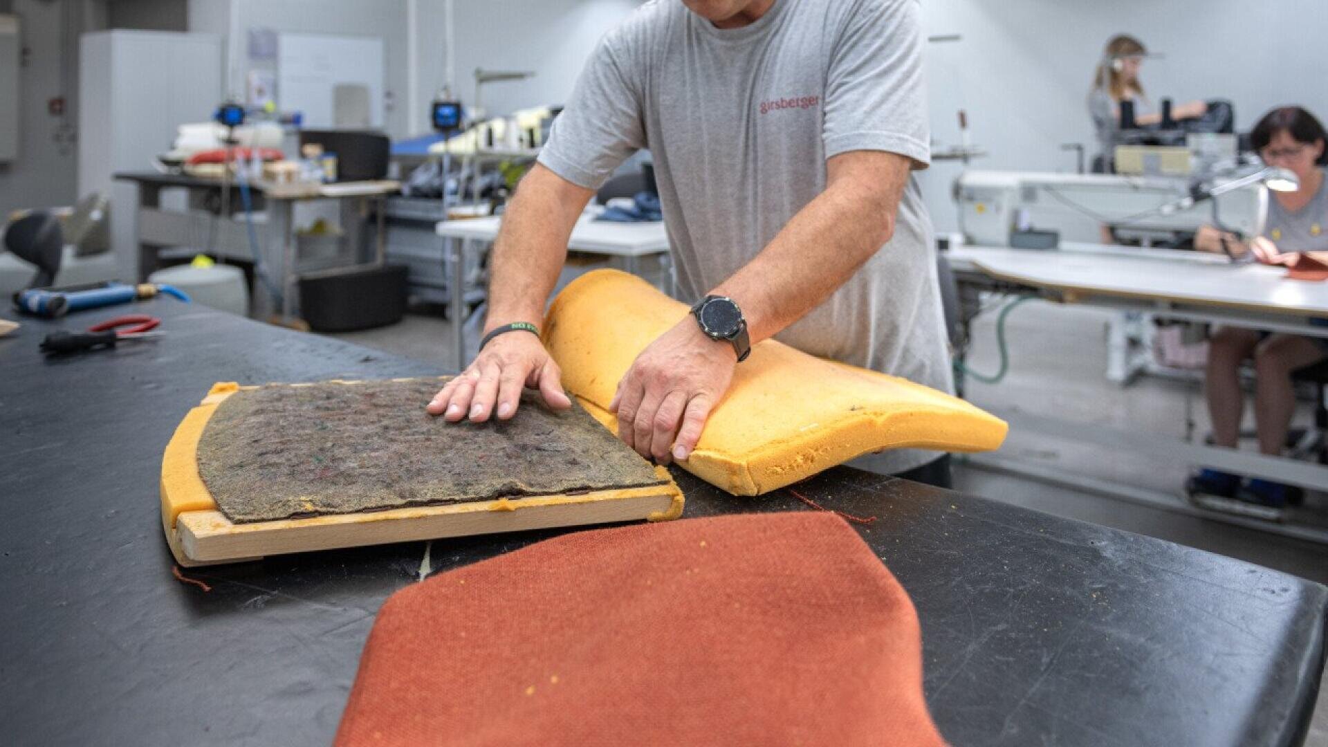 Une personne vêtue d'une chemise grise assemble un coussin de chaise en pressant de la mousse sur une base d'assise à une table de travail dans un atelier. Des machines à coudre et d'autres travailleurs sont visibles à l'arrière-plan.