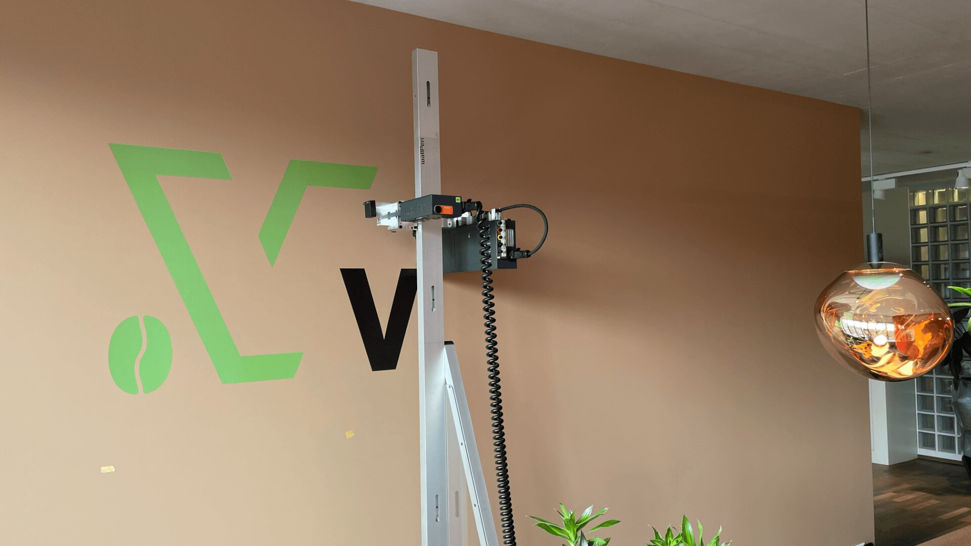 Ein Wanddrucker druckt ein grünes und schwarzes Logo auf eine beigefarbene Wand in einem modernen Raum, in dessen Nähe eine bernsteinfarbene Hängelampe und grüne Pflanzen zu sehen sind.