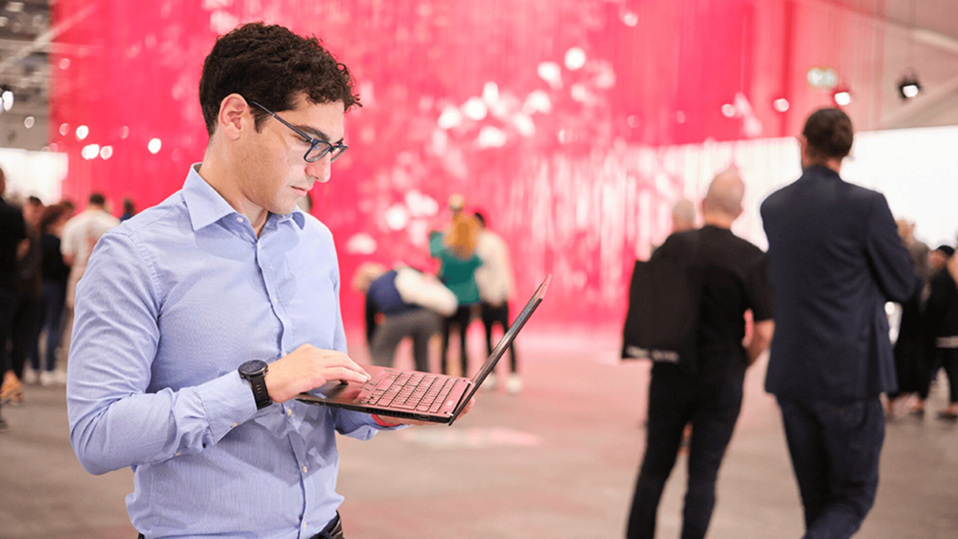Ein Mann mit blauem Hemd und Brille tippt auf einem Laptop in einem hellen Innenraum mit rosa Hängedekorationen, während im unscharfen Hintergrund mehrere Menschen herumlaufen und sich versammeln.