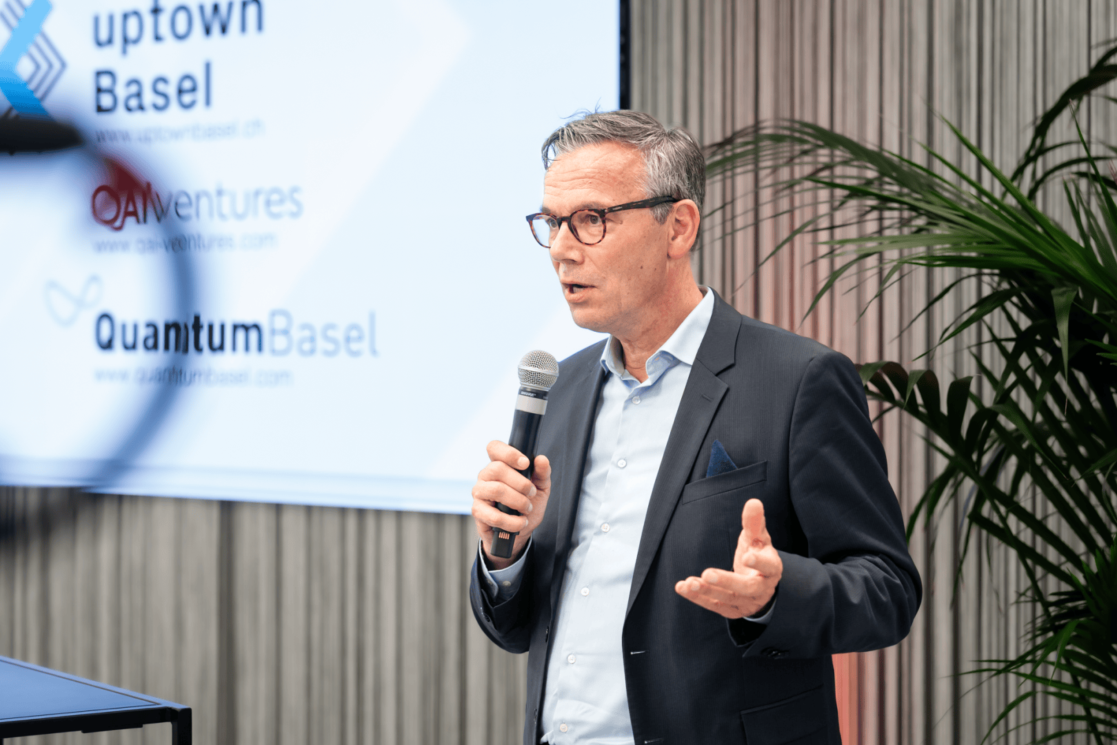 Ein Mann im Anzug und mit Brille spricht bei einer Veranstaltung in ein Mikrofon. Hinter ihm ist ein Bildschirm mit den Logos von Uptown Basel, QAi Ventures und Quantum Basel zu sehen, im Hintergrund sind Pflanzen und vertikale Jalousien zu sehen.