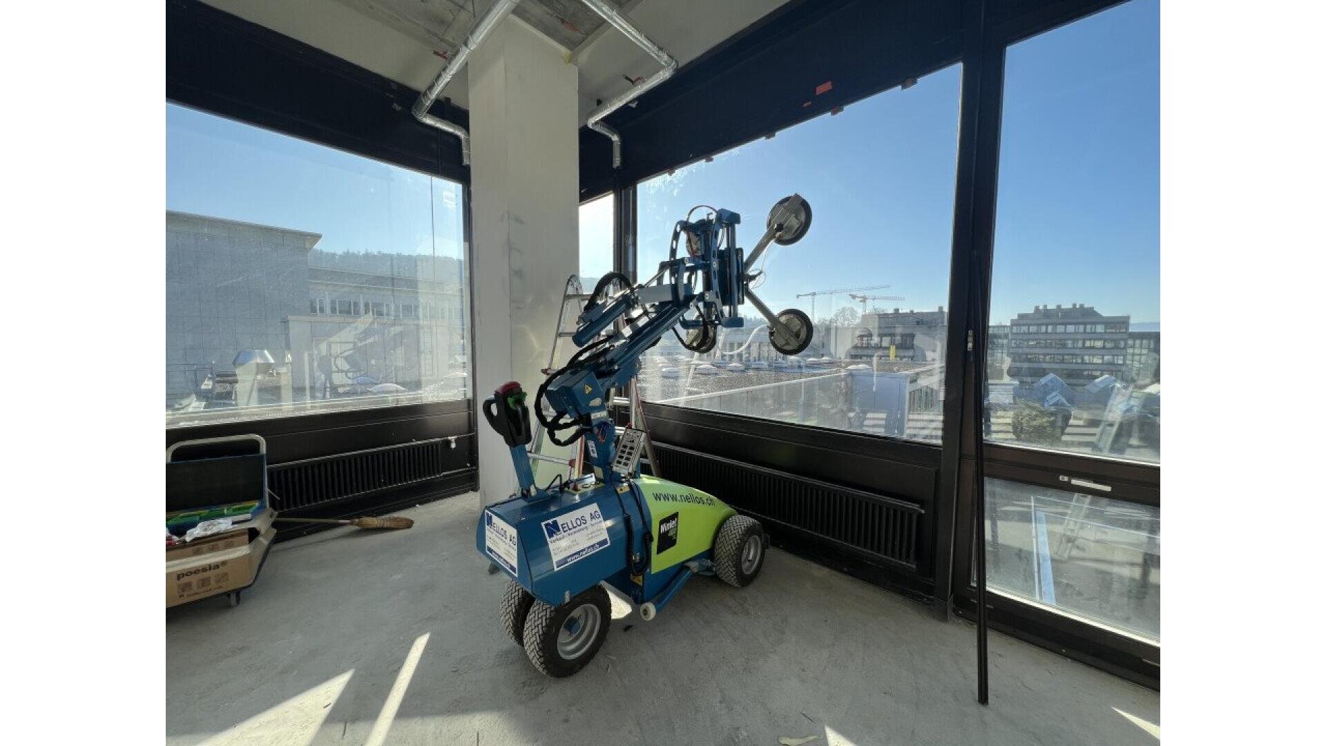 Un robot d'installation en verre muni de ventouses se trouve dans l'angle d'un immeuble de bureaux moderne en construction, entouré de grandes fenêtres et d'équipements de construction.