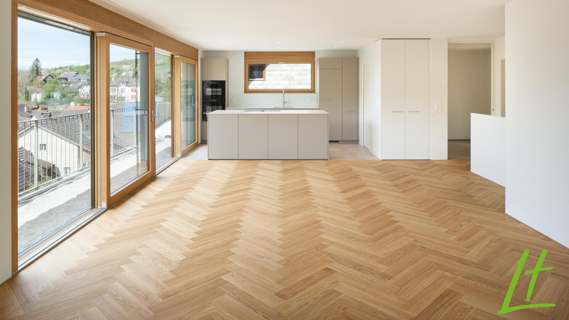 Appartement moderne, non meublé, avec de grandes fenêtres, un sol en bois à chevrons, une cuisine ouverte avec des armoires blanches, et une vue sur les toits et la verdure à l'extérieur. Logo vert "LT" dans le coin inférieur droit.