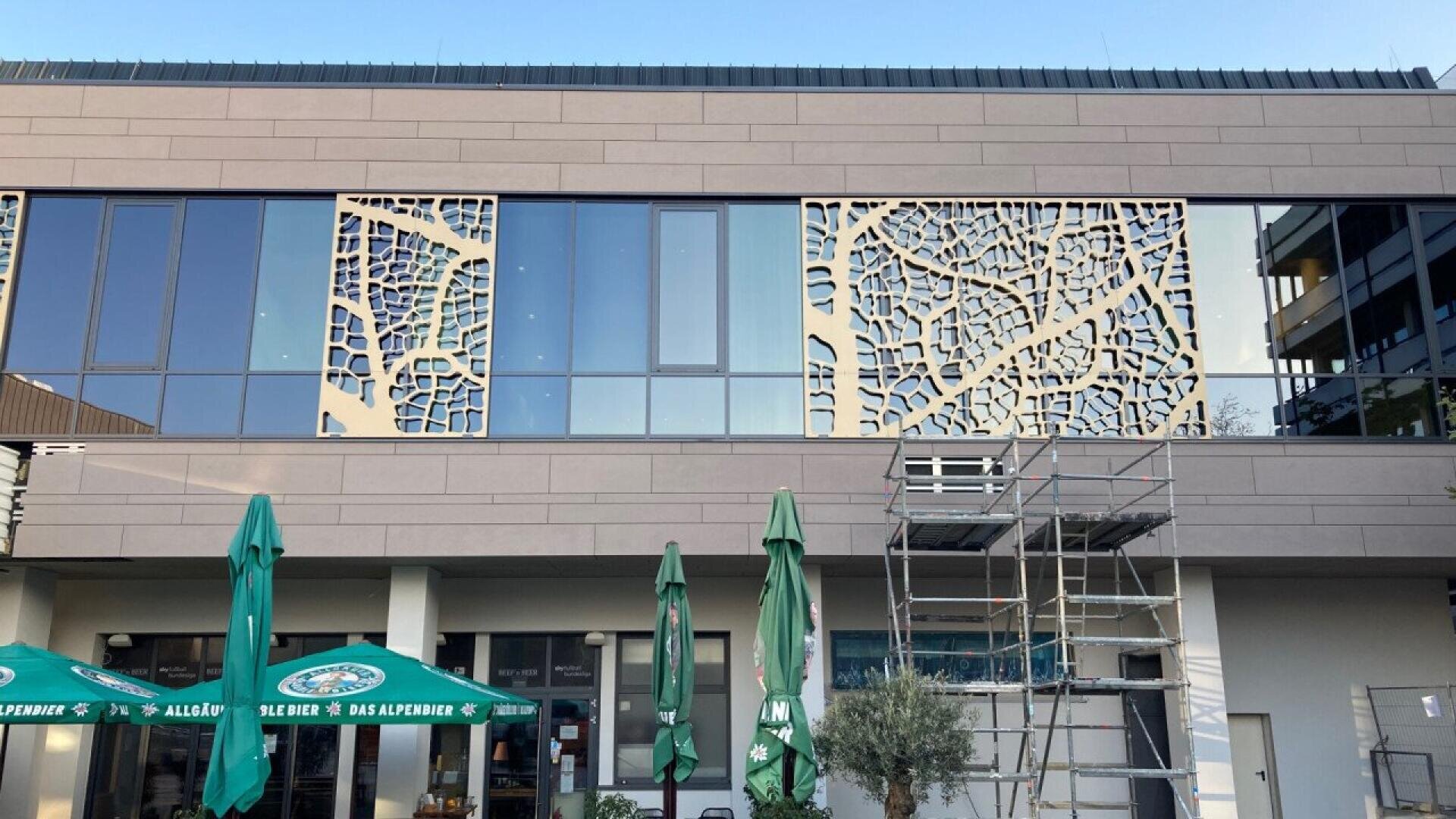 Façade d'un bâtiment moderne avec des panneaux décoratifs, des fenêtres en verre et une structure d'échafaudage à l'avant. Plusieurs parasols verts sont ouverts près de l'entrée au rez-de-chaussée.