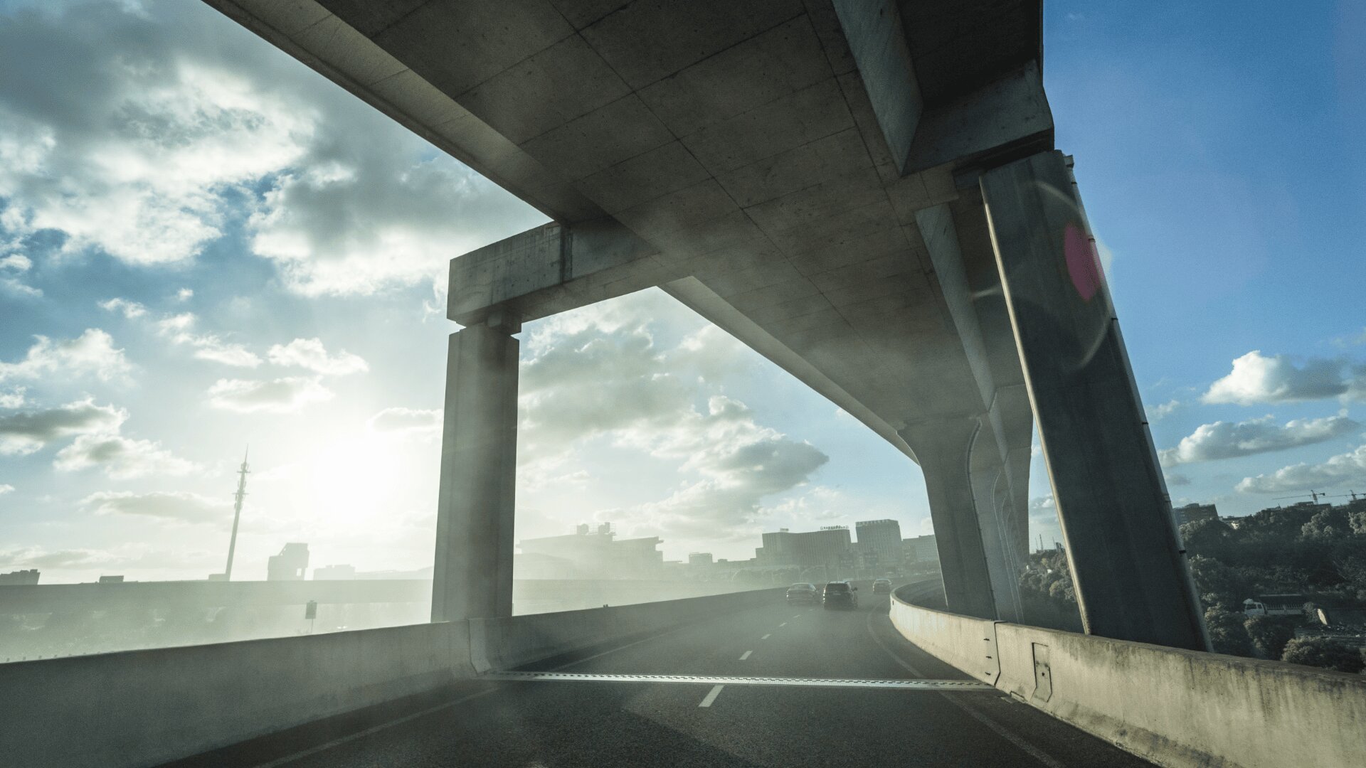 Eine Autobahnüberführung mit großen Betonpfeilern ragt über eine sonnenbeschienene Straße, unter der Autos fahren. Der Himmel ist teilweise bewölkt, und im Hintergrund ist die Skyline einer Stadt zu sehen.