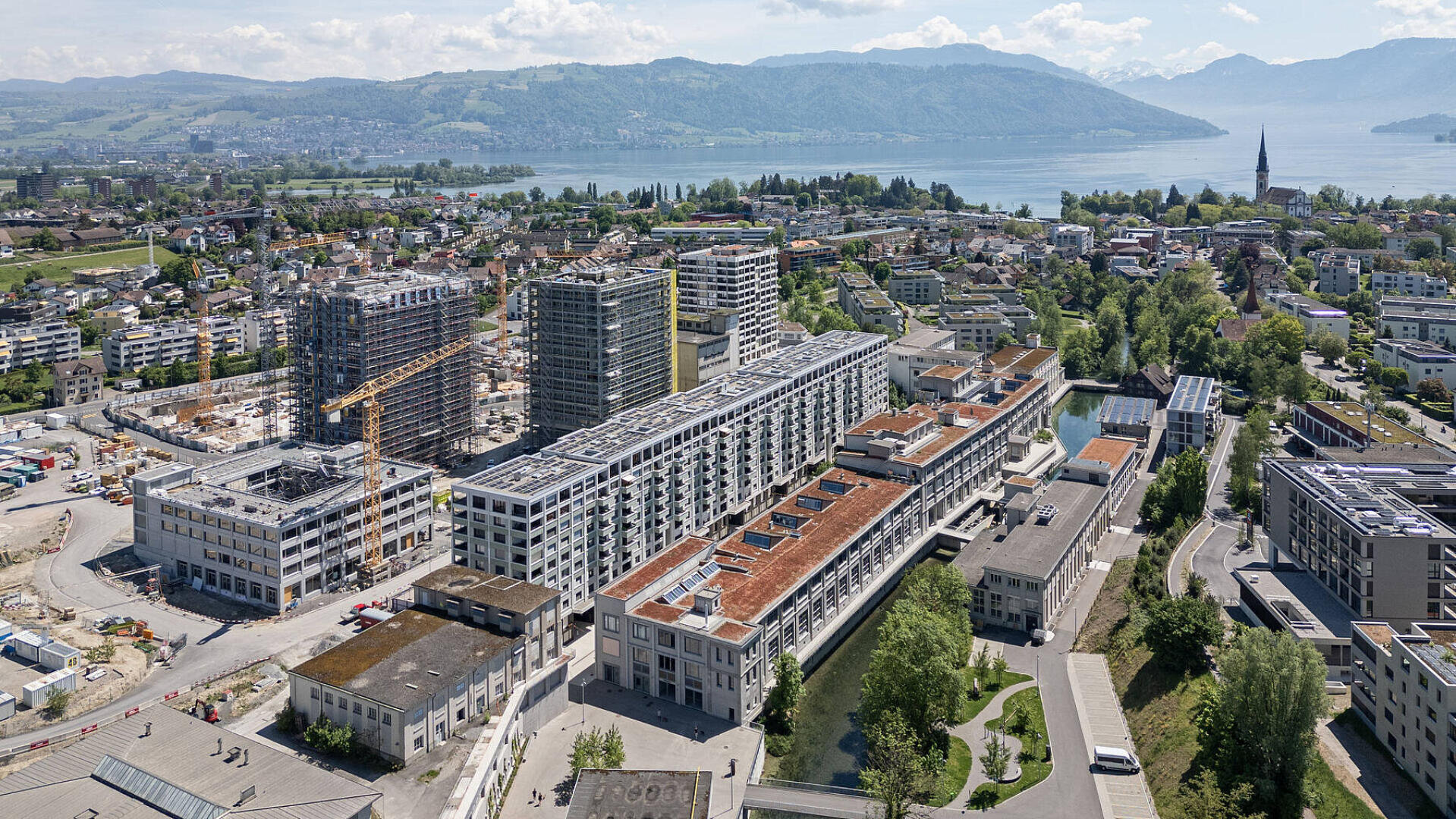 Vue aérienne d'une ville moderne avec des immeubles de moyenne hauteur, des grues de construction et un lac en arrière-plan. Des montagnes et de la verdure entourent la zone, et un clocher d'église est visible parmi les maisons.