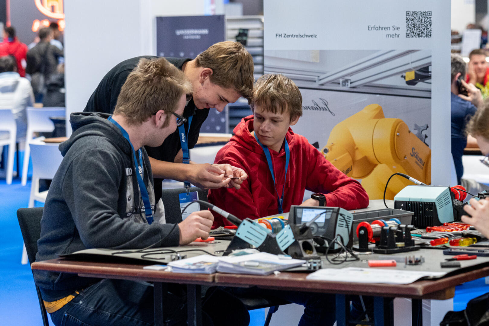 Drei junge Leute sitzen gemeinsam an einem Tisch mit Elektronikgeräten und untersuchen ein kleines Bauteil. Sie wirken engagiert und konzentriert, während vor ihnen verschiedene Geräte, Kabel und Werkzeuge ausgebreitet sind.