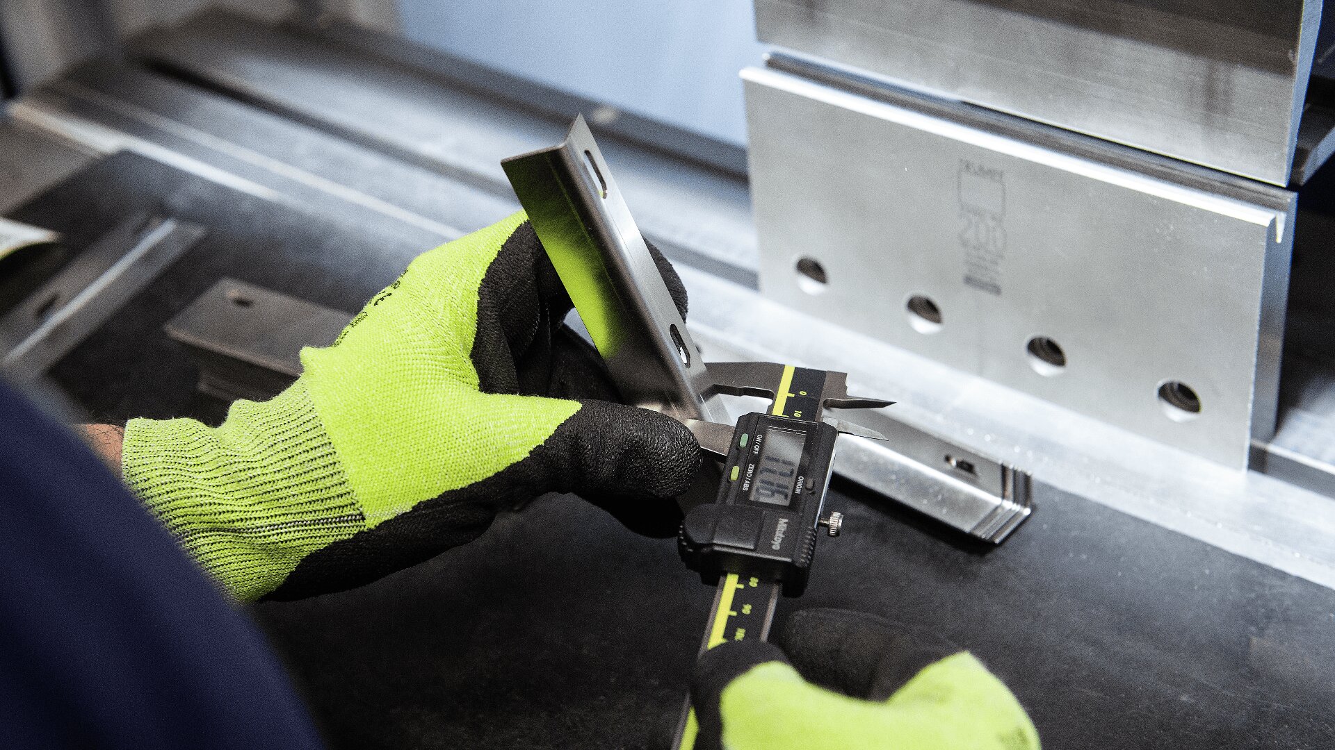Eine Person mit leuchtend grünen Handschuhen misst mit einem digitalen Messschieber ein Metallteil in einer Werkstatt, wobei im Hintergrund Maschinen und andere Metallteile zu sehen sind.