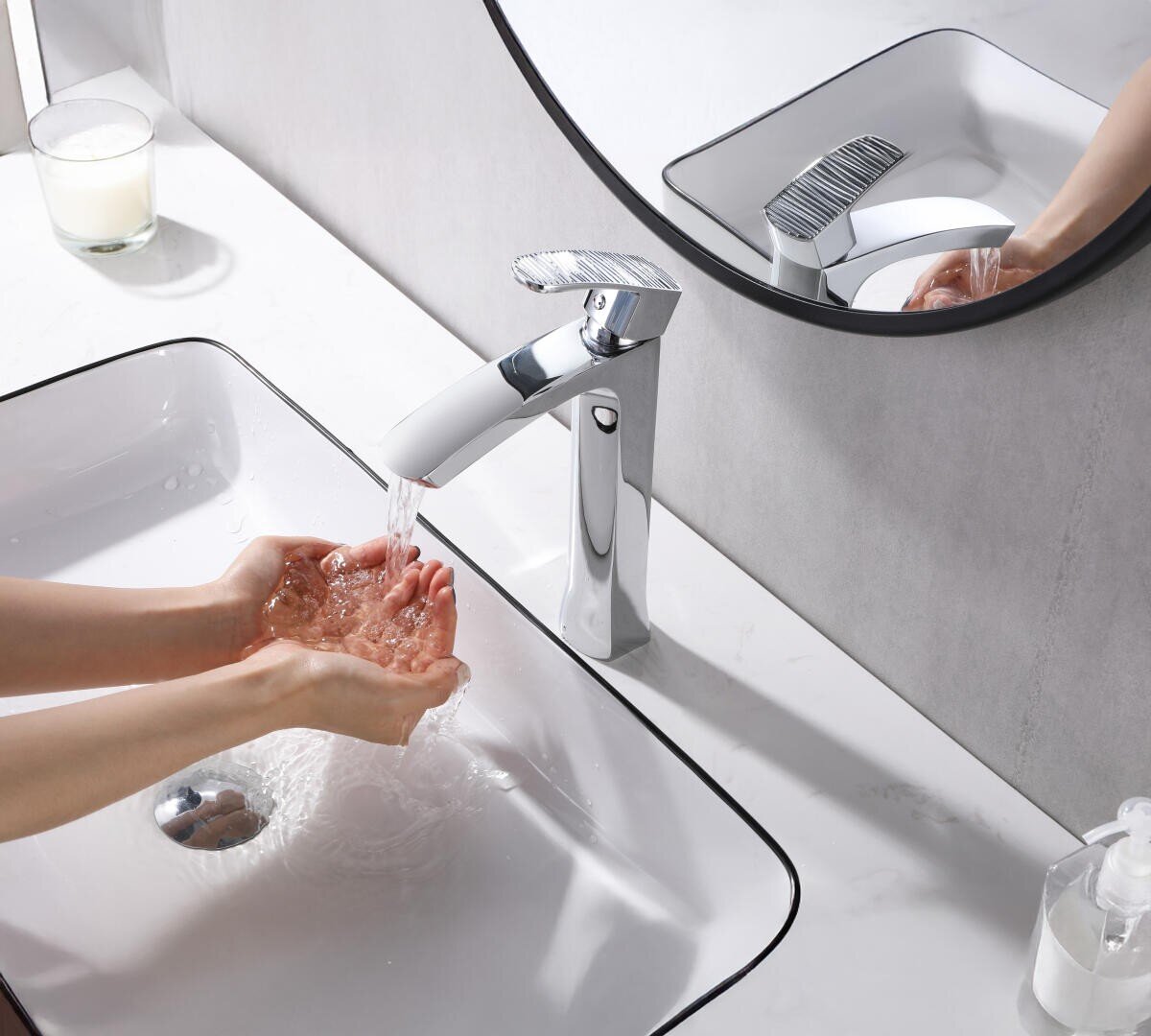 Une personne se lave les mains sous un robinet moderne chromé dans un lavabo blanc, faisant gicler l'eau. Un miroir rond, un distributeur de savon et une bougie en verre sont posés sur le plan de travail blanc.