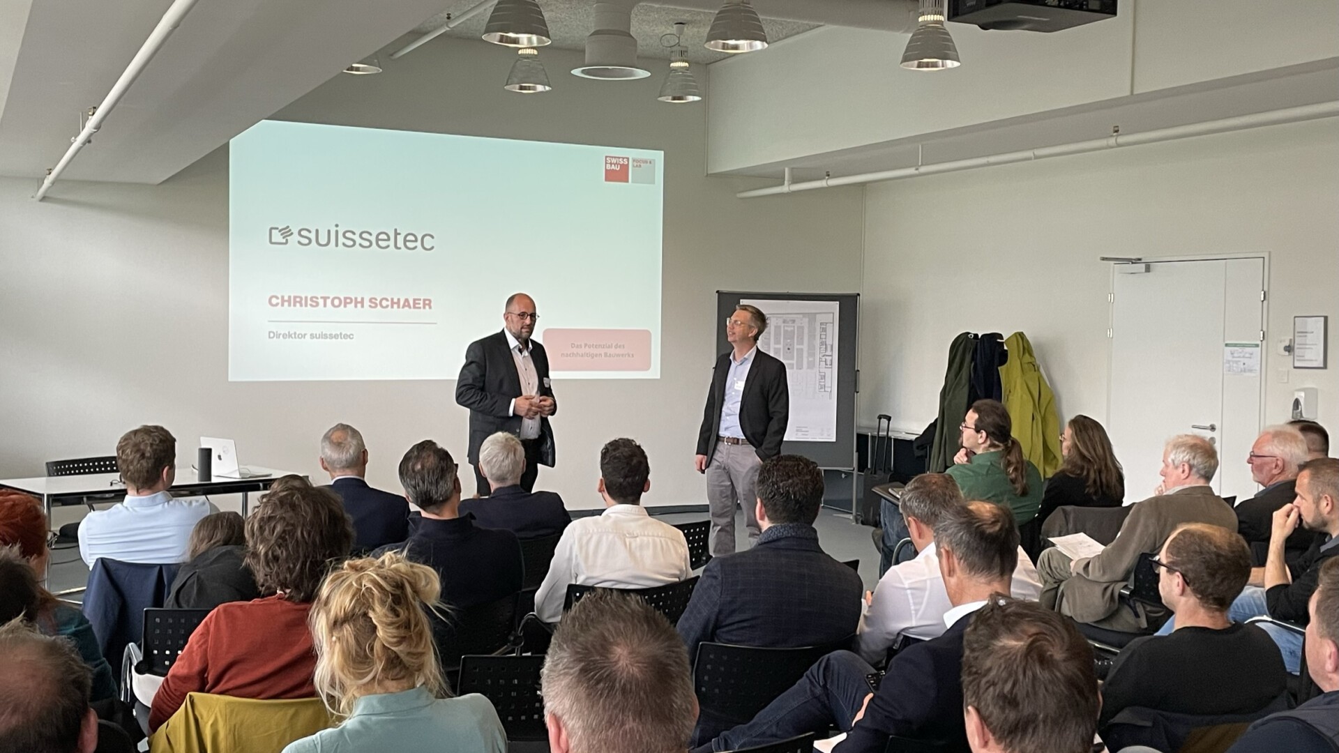 Ein Mann im Anzug spricht in einem Konferenzraum zu seinem Publikum. Hinter ihm ist eine Präsentationsfolie zu sehen, auf der suissetec und Christoph Schaer zu sehen sind. Ein weiterer Mann steht neben ihm, und die Anwesenden hören aufmerksam zu.