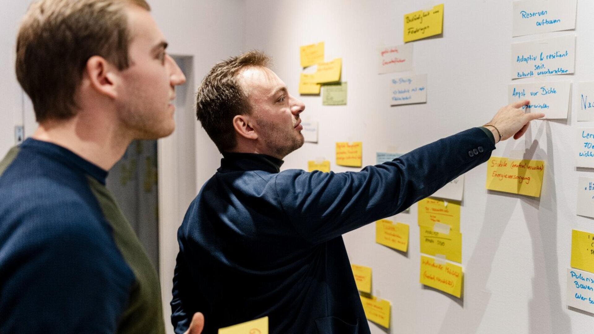 Deux hommes se tiennent devant un mur couvert de notes adhésives colorées. L'un d'eux pointe une note tandis que l'autre l'observe, ce qui suggère une séance de brainstorming ou de planification en collaboration.