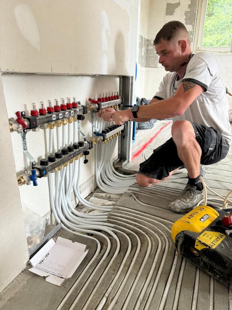 Ein Klempner kniet auf dem Boden und arbeitet an einem komplexen System aus weißen Rohren und Ventilen, die aus einer Wand herausragen. Werkzeuge und Papiere liegen in der Nähe, und der Raum scheint sich im Bau oder in der Renovierung zu befinden.