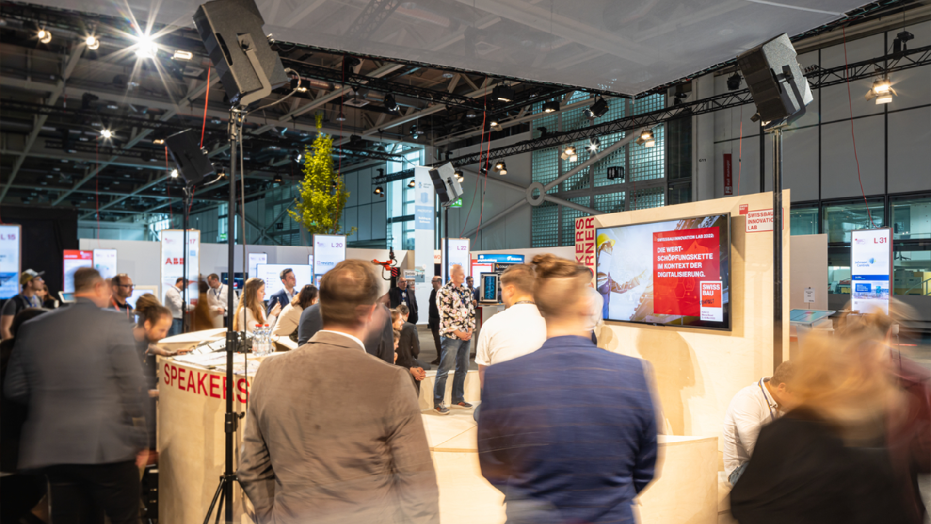 Eine Gruppe von Personen nimmt an einer Präsentation oder einem Vortrag bei einer modernen Indoor-Veranstaltung oder Konferenz teil, einige sitzen, andere stehen. Große Bildschirme und Banner sind zu sehen, und die Umgebung wirkt geschäftig und professionell.