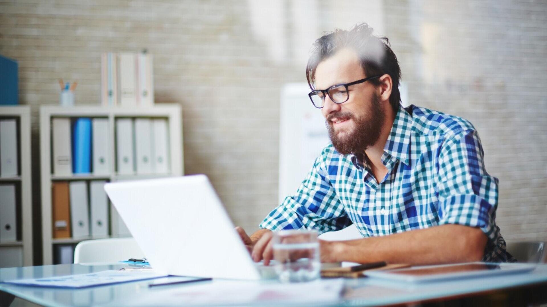 Un homme barbu et portant des lunettes, vêtu d'une chemise bleue à carreaux, est assis à un bureau et travaille sur un ordinateur portable dans un bureau moderne. Des classeurs, des documents et un verre d'eau sont posés sur le bureau.