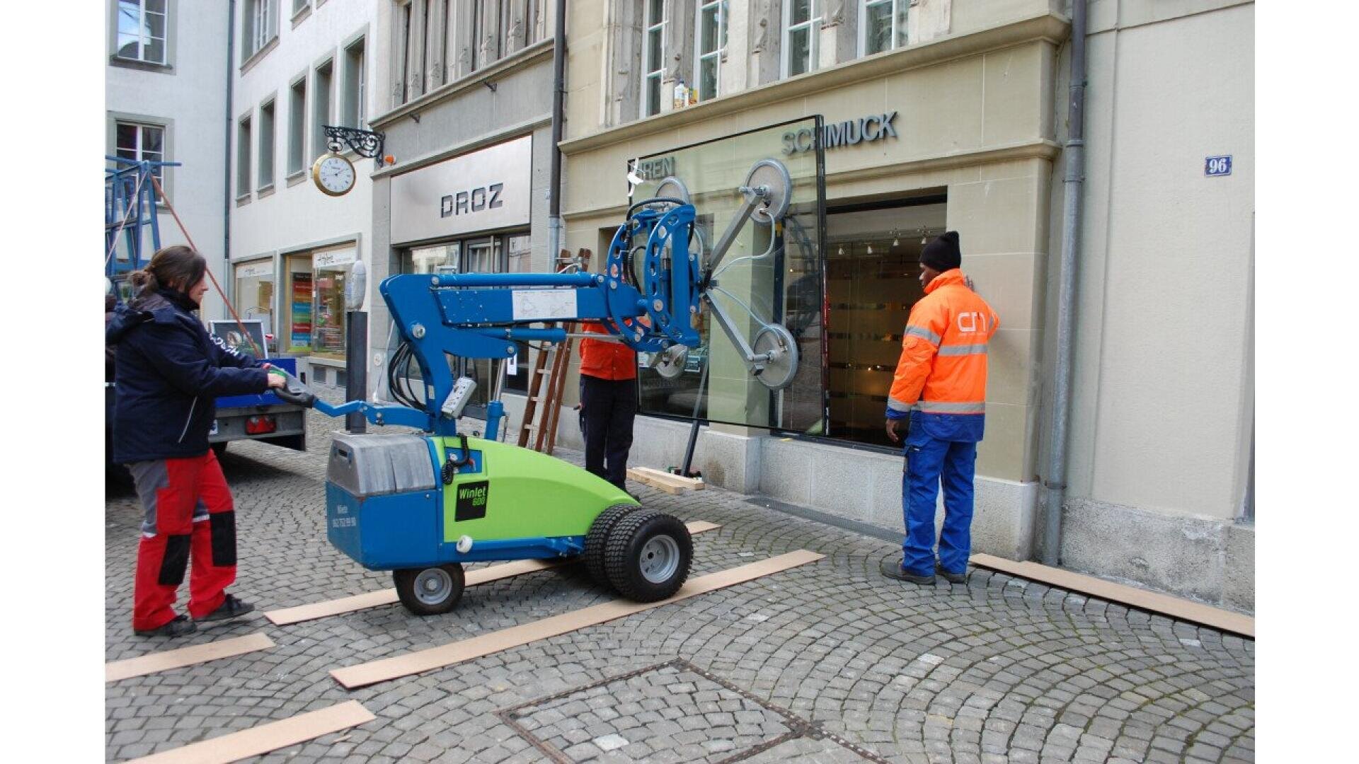 Trois ouvriers utilisent une machine à lever le verre bleue et verte pour installer une grande vitre dans la devanture d'un magasin situé dans une rue pavée. Deux des travailleurs portent des gilets de sécurité orange.