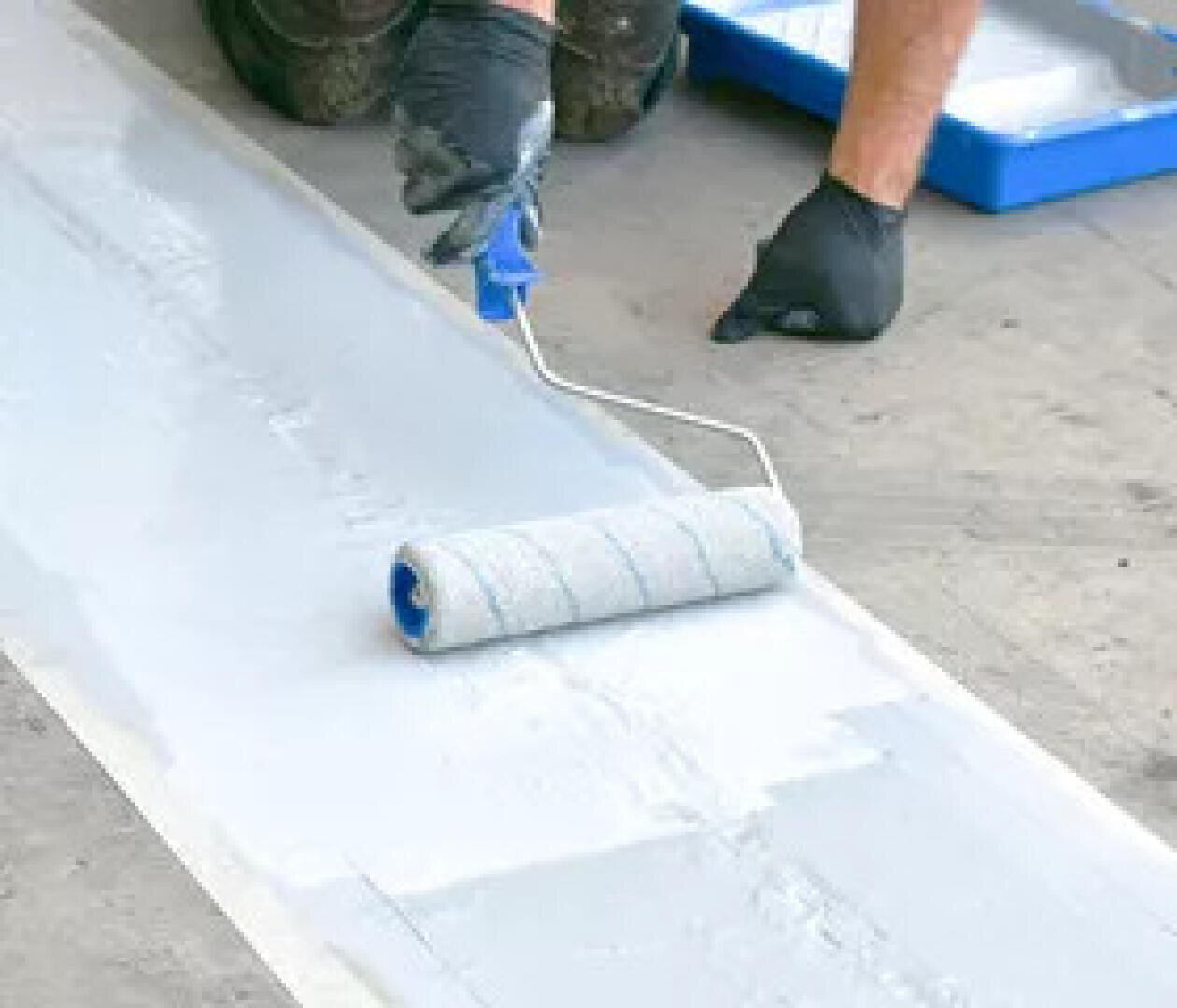 Une personne portant des gants noirs applique de la peinture blanche sur une bande de sol à l'aide d'un rouleau à peinture, avec un bac à peinture visible en arrière-plan.