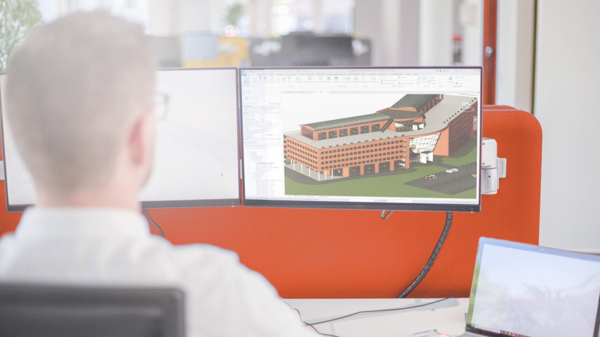 Une personne est assise à un bureau où se trouvent deux écrans d'ordinateur ; l'un d'eux affiche un modèle architectural en 3D d'un bâtiment, tandis que l'autre est essentiellement blanc. Un ordinateur portable est également ouvert sur le bureau.