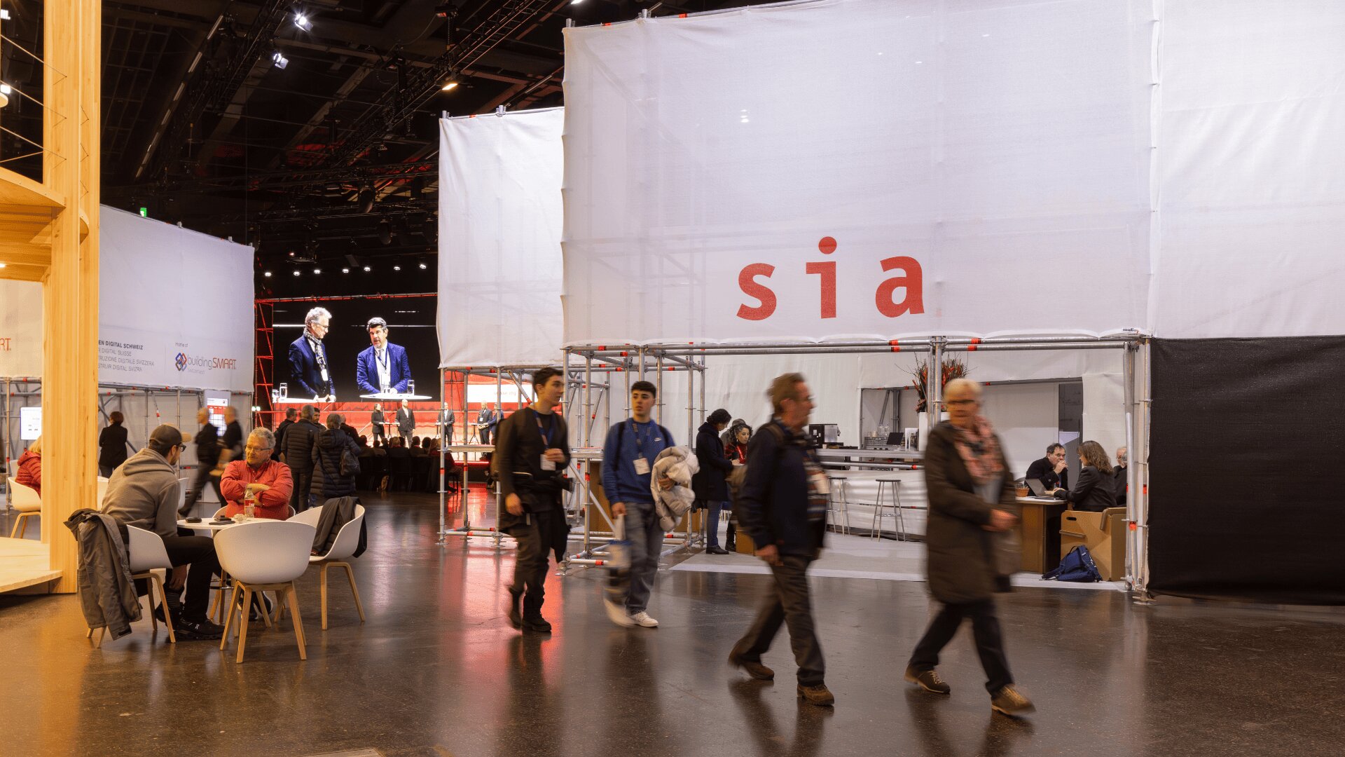 Menschen gehen und sitzen in einer belebten Ausstellungshalle mit weißen Ständen, Sitzgelegenheiten und einem großen Schild mit der Aufschrift sia. Ein Bildschirm im Hintergrund zeigt zwei Personen, die auf der Bühne sprechen.