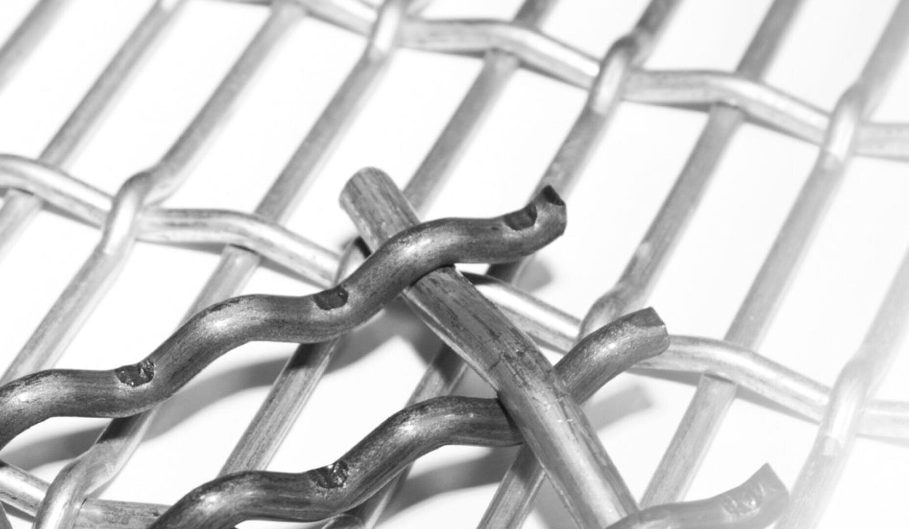 Vue rapprochée de fils métalliques entrelacés formant une grille, avec deux tiges métalliques ondulées placées en diagonale au-dessus de la grille. L'image est en niveaux de gris.