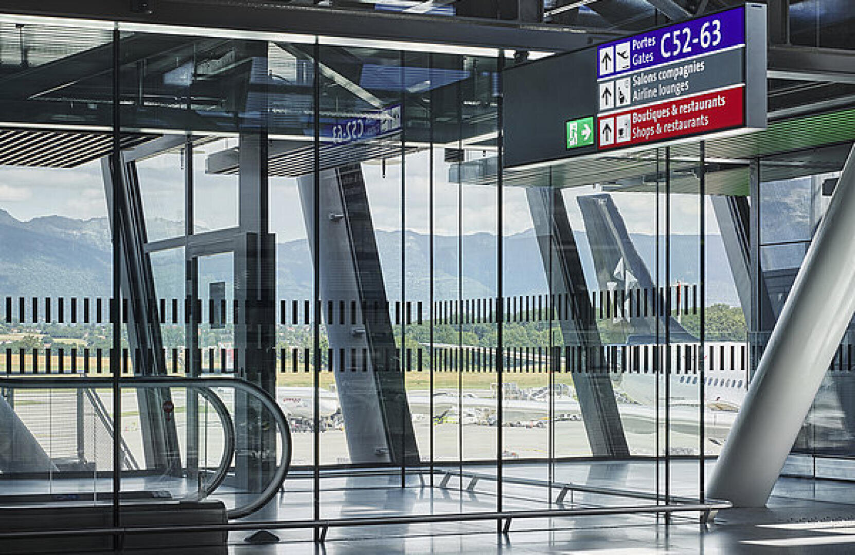 Modernes Flughafenterminal mit großen Glasfenstern, die einen Blick auf die Start- und Landebahn und die umliegenden Berge bieten. Überkopfschilder zeigen in mehreren Sprachen den Weg zu den Gates, Geschäften und Restaurants an.