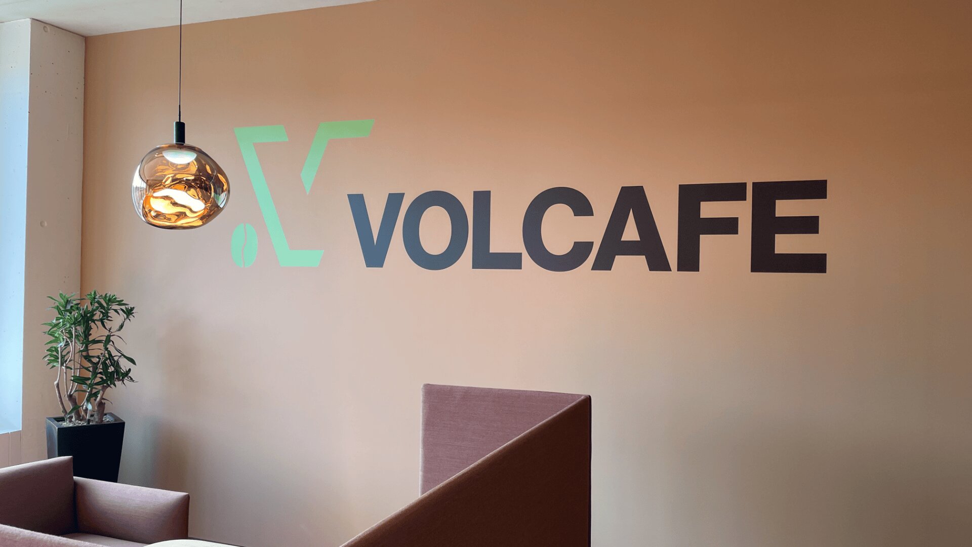 Ein moderner Büroraum mit dem Wort VOLCAFE und einem grünen Logo an einer pfirsichfarbenen Wand, einer hängenden Glasleuchte, einer kleinen Pflanze und rosafarbenen Sitzkabinen.