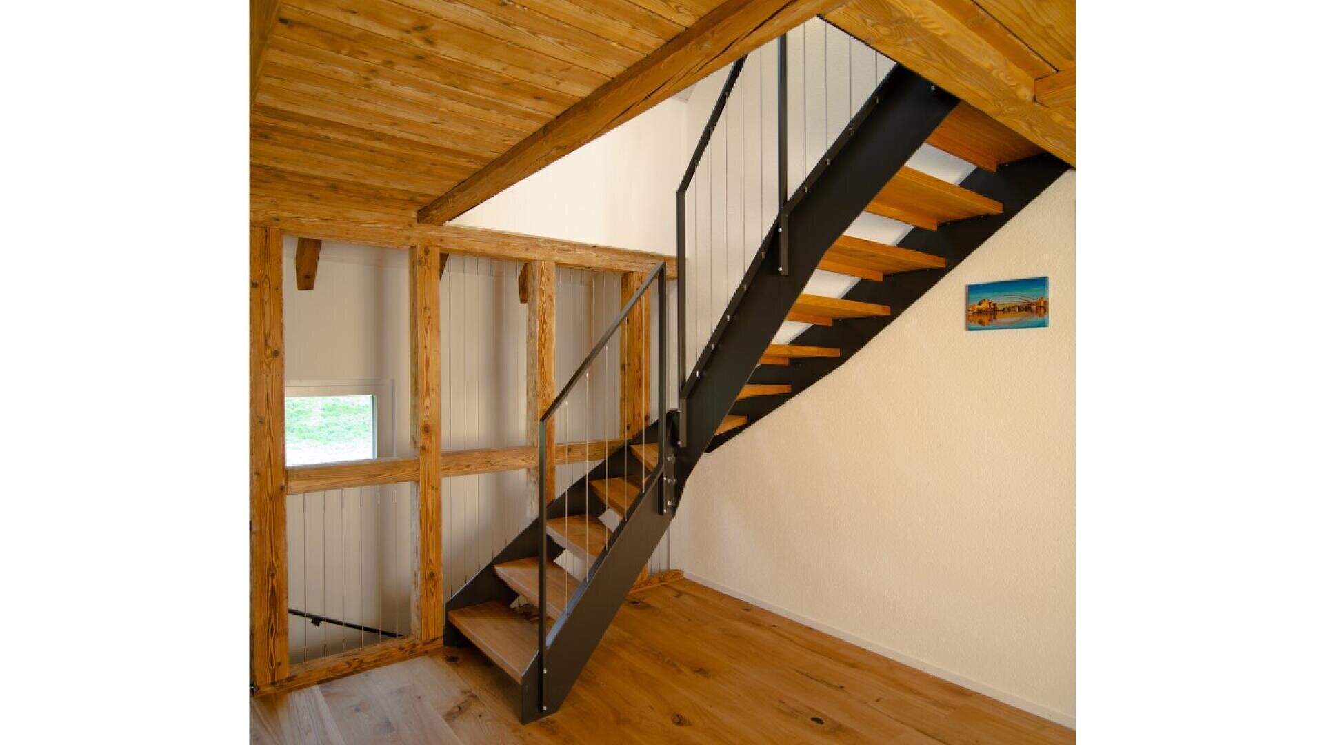 Un escalier moderne en bois avec des rampes en métal noir mène à une pièce lumineuse avec des planchers et un plafond en bois. Une petite fenêtre et un tableau de paysage sont accrochés aux murs de couleur crème.