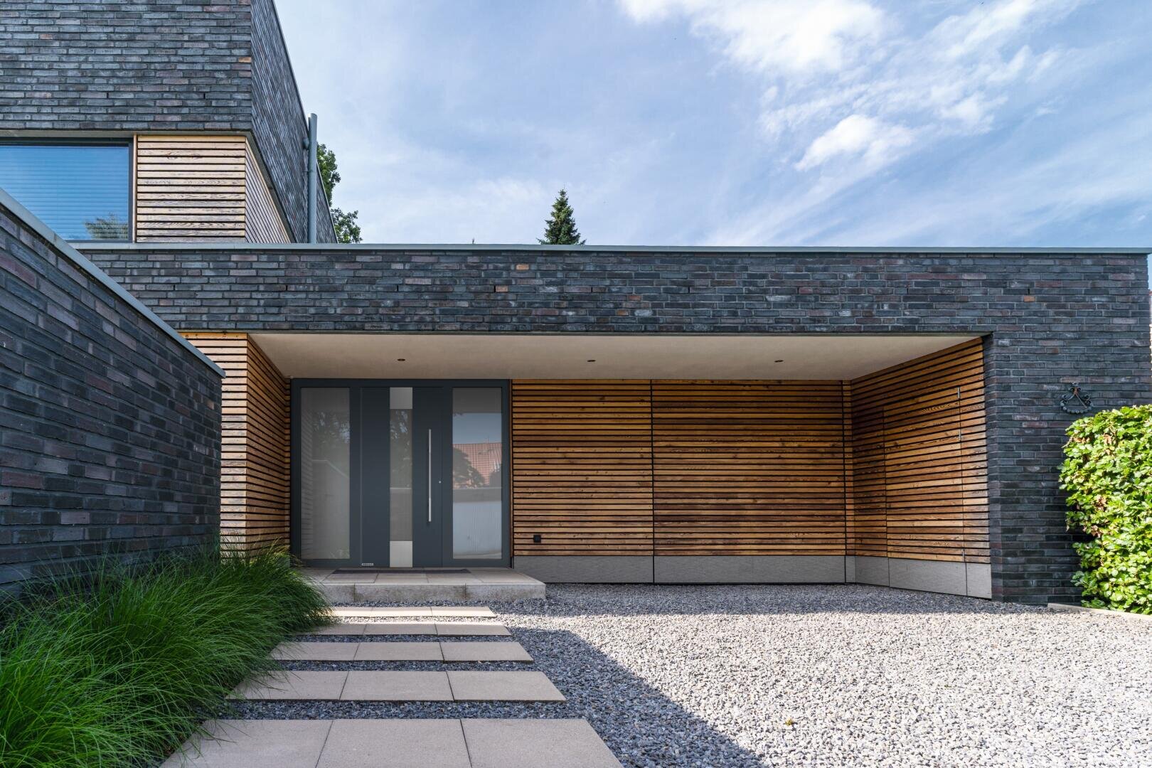 Modernes Haus mit dunklem Backstein und horizontalen Holzlatten, einer großen Glastür, einem Hof aus Kies, einem steinernen Gehweg und gepflegter Bepflanzung entlang der linken und rechten Seite.