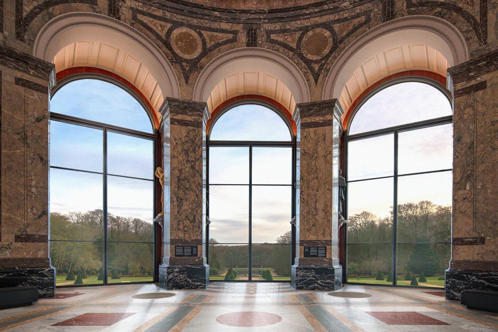 Große, von Marmorsäulen eingerahmte Bogenfenster geben den Blick frei auf einen malerischen Garten mit Bäumen und gepflegten Rasenflächen, der von einer eleganten, großen Halle mit gemustertem Steinboden und hoher Decke aus gesehen wird.