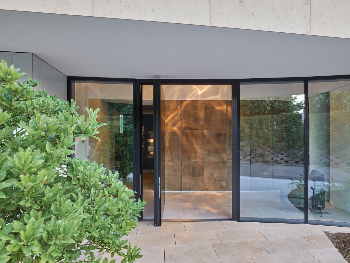 Une entrée de bâtiment moderne avec de grandes portes et fenêtres vitrées, un sol en pierre claire et une plante à feuilles vertes au premier plan. L'intérieur est décoré de boiseries chaudes et d'un éclairage doux.