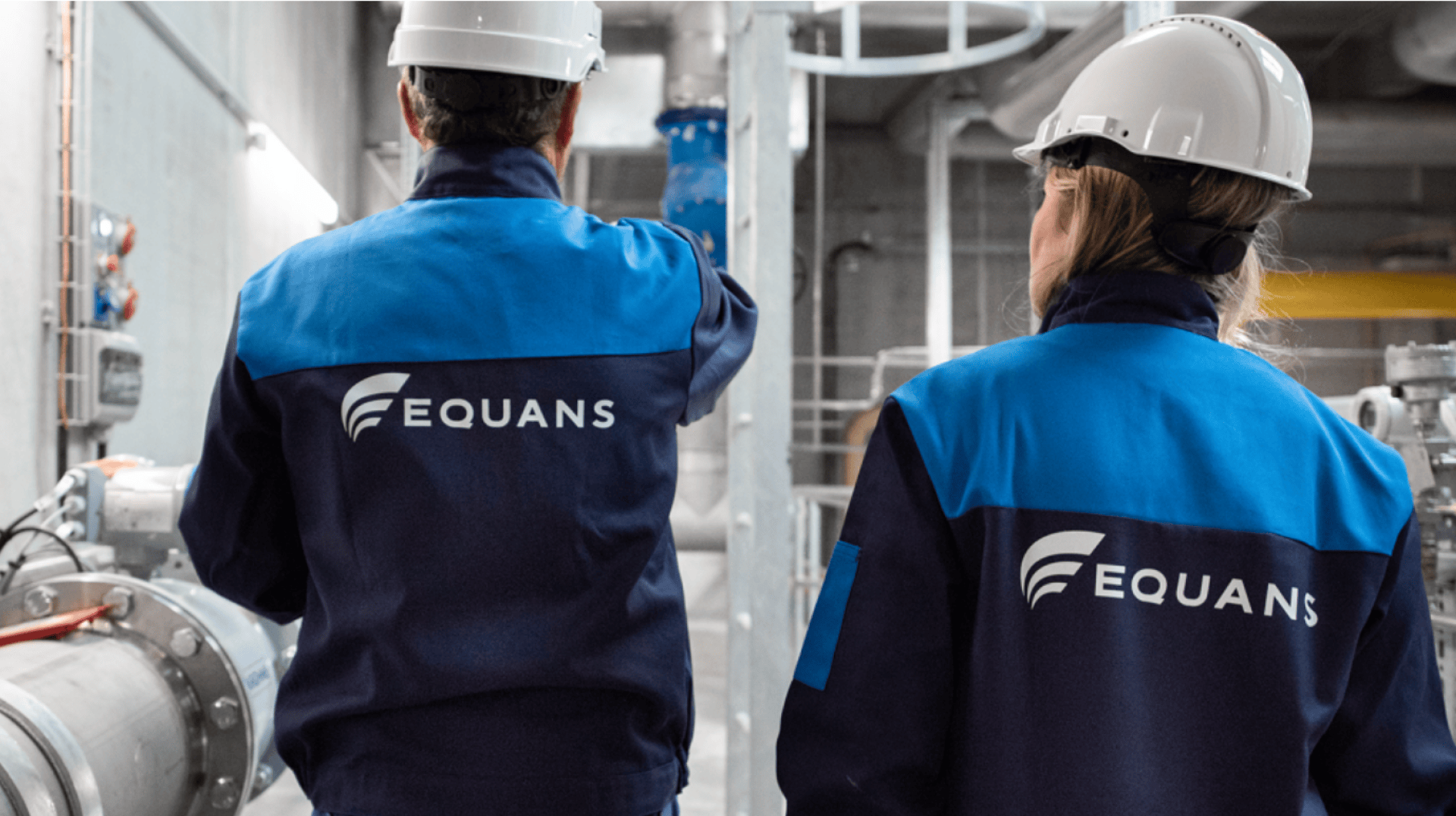 Deux personnes portant des casques de sécurité blancs et des uniformes bleus avec le logo EQUANS sont debout dans une installation industrielle, faisant face à l'extérieur et observant l'équipement et la tuyauterie.