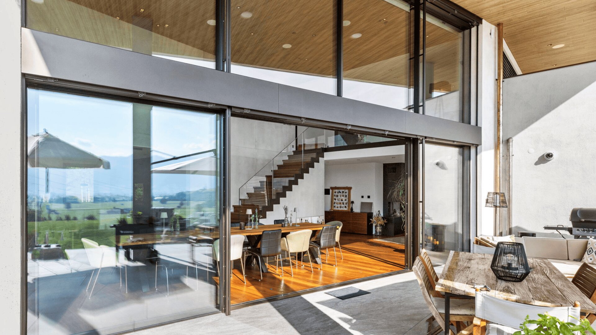 Modernes Haus mit großen Glasschiebetüren, die zu einem hellen Essbereich mit Holzböden und einer Treppe führen. Die Außenterrasse verfügt über einen Holztisch, Stühle und Blick auf grüne Felder und Berge.