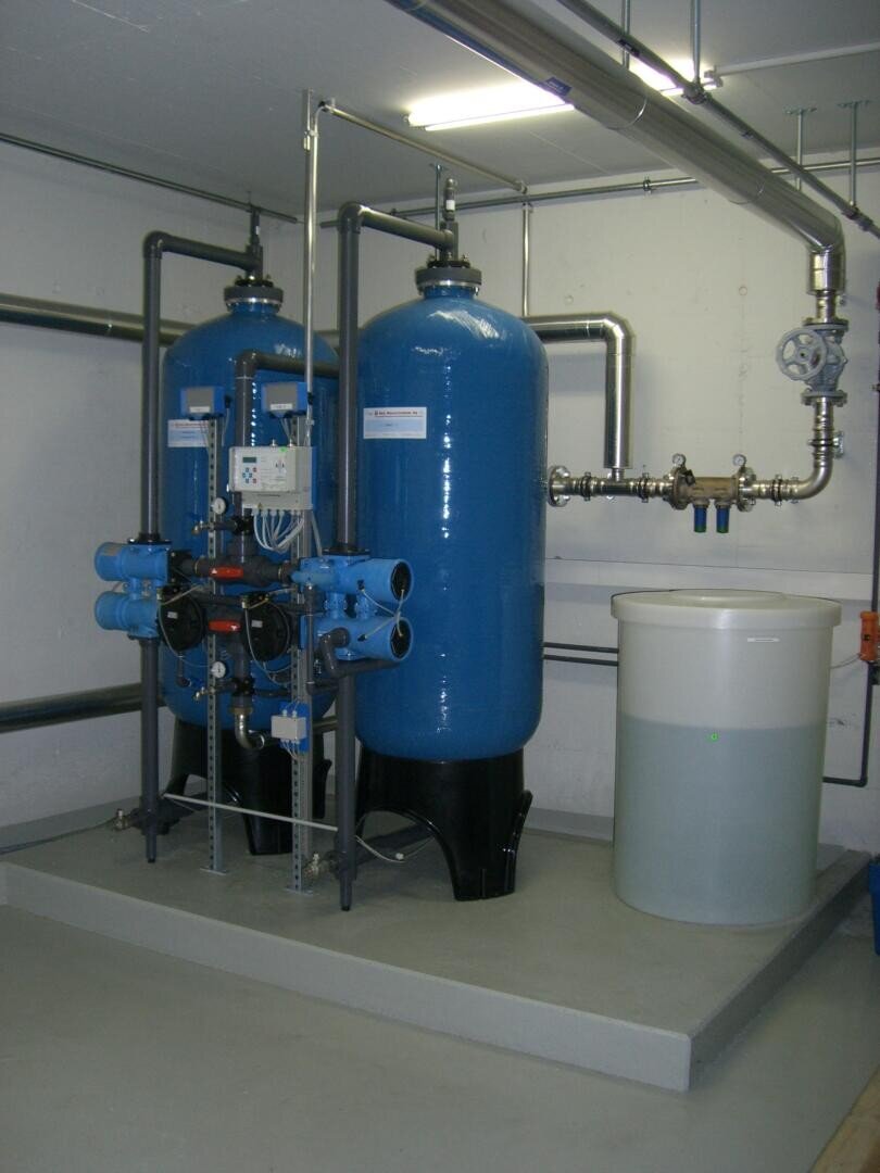 Zwei große blaue industrielle Wasserfiltertanks, die durch Rohre miteinander verbunden sind, stehen auf einer Plattform in einem sauberen, weißen Raum, daneben ein weißer Plastikbehälter, der teilweise mit Wasser gefüllt ist.