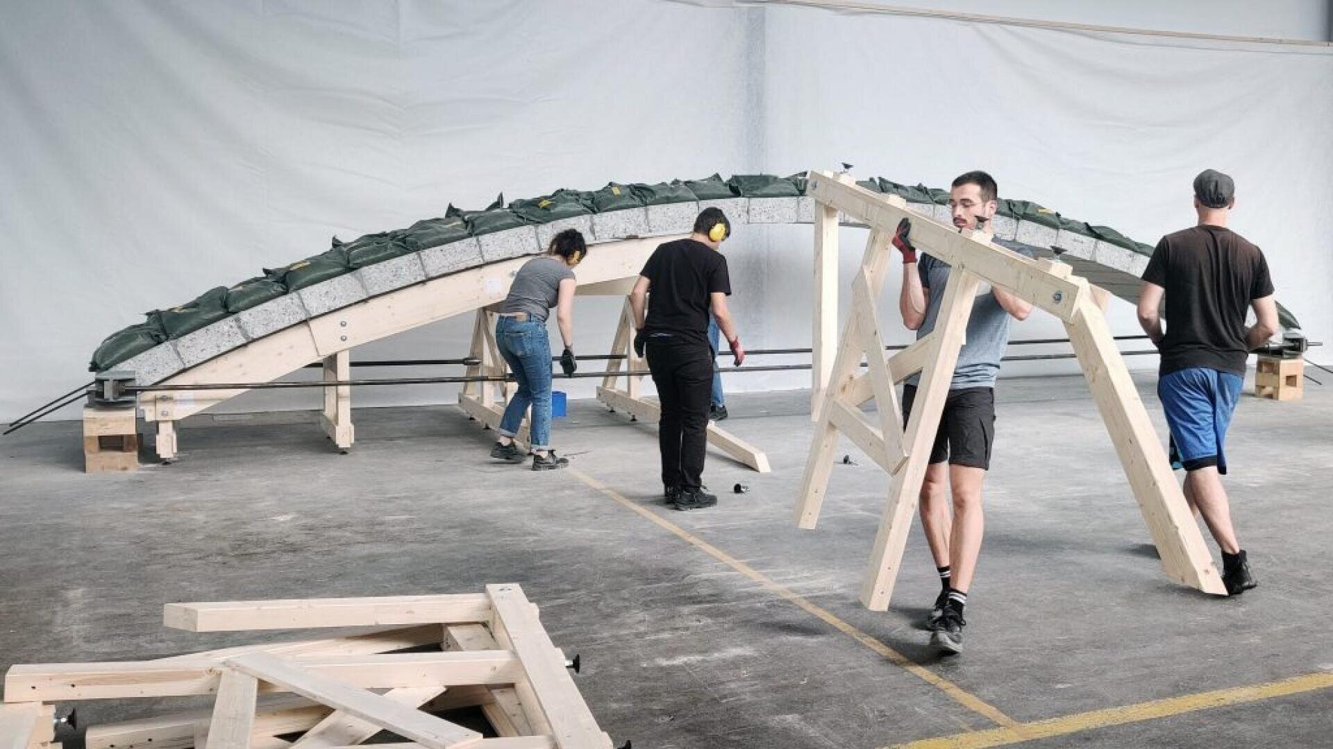 Vier Personen bauen im Haus eine große bogenförmige Holzstruktur zusammen. Eine Person trägt einen Holzrahmen, während die anderen an verschiedenen Teilen des Bogens arbeiten, der von Balken gestützt und mit grünen Säcken beschwert wird.