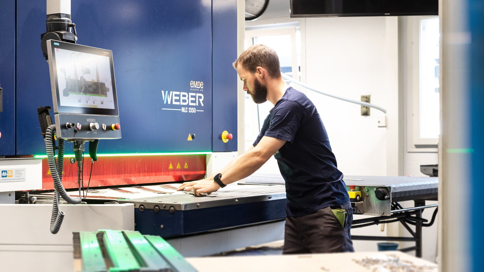 Ein Mann in einem marineblauen Hemd bedient eine große blaue Weber-CNC-Maschine in einem hellen Industriearbeitsraum und konzentriert sich auf die Einstellung von Materialien auf der Oberfläche der Maschine.