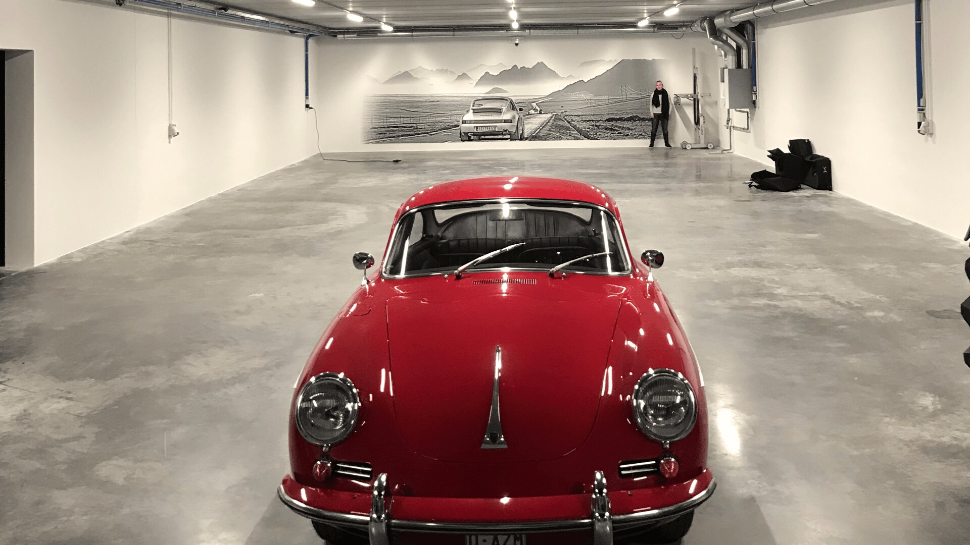 Ein leuchtend roter Oldtimer steht in einer geräumigen, leeren Garage mit weißen Wänden und einem polierten Betonboden. Ein großes Wandgemälde mit einem Oldtimer und Bergen ziert die Rückwand, daneben steht eine Person.