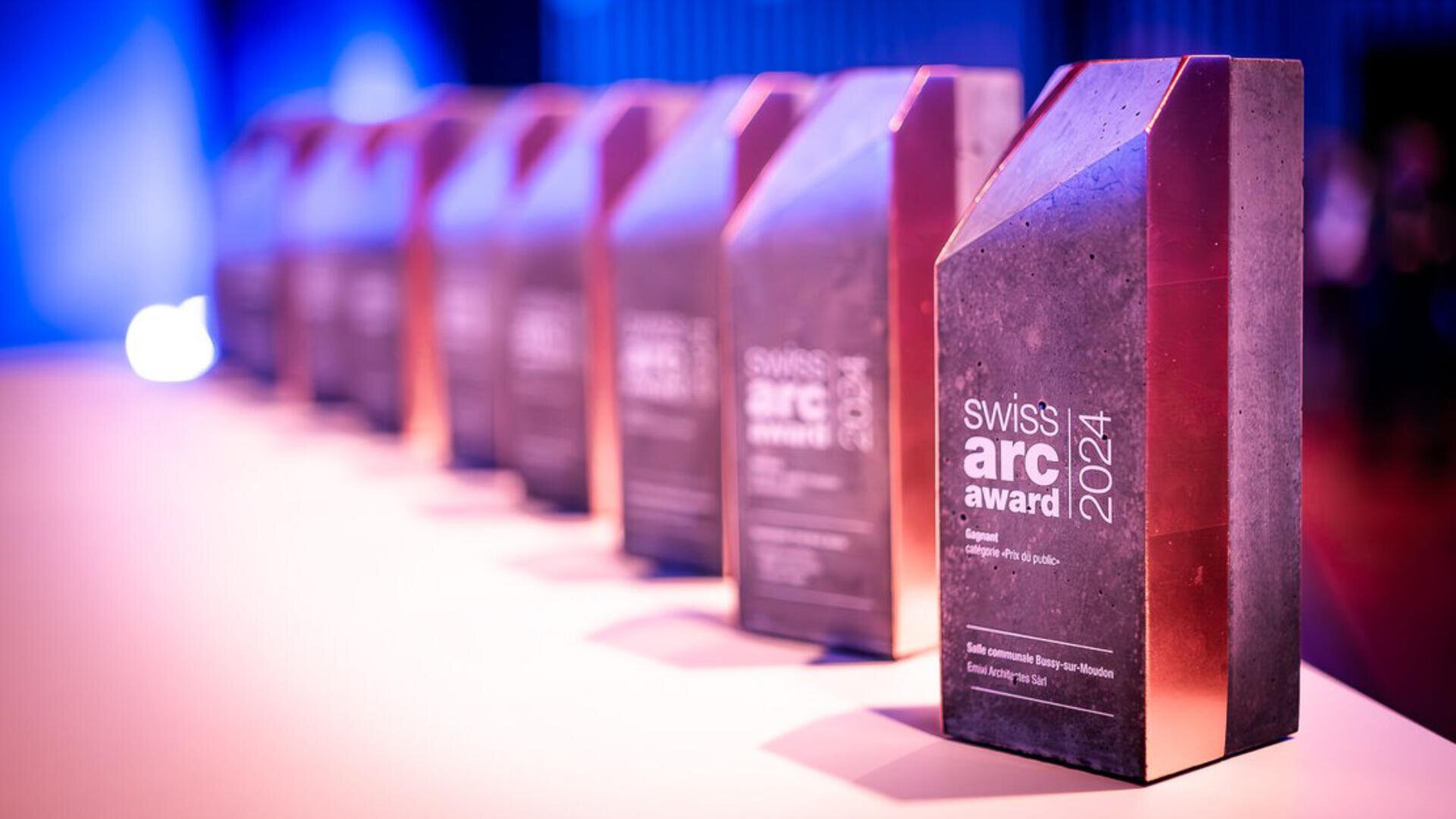 Eine Reihe von Swiss Arc Award 2024 Trophäen aus Metall und Beton sind auf einem Tisch in einer hell erleuchteten Umgebung mit einem lila und blauen Hintergrund aufgereiht.