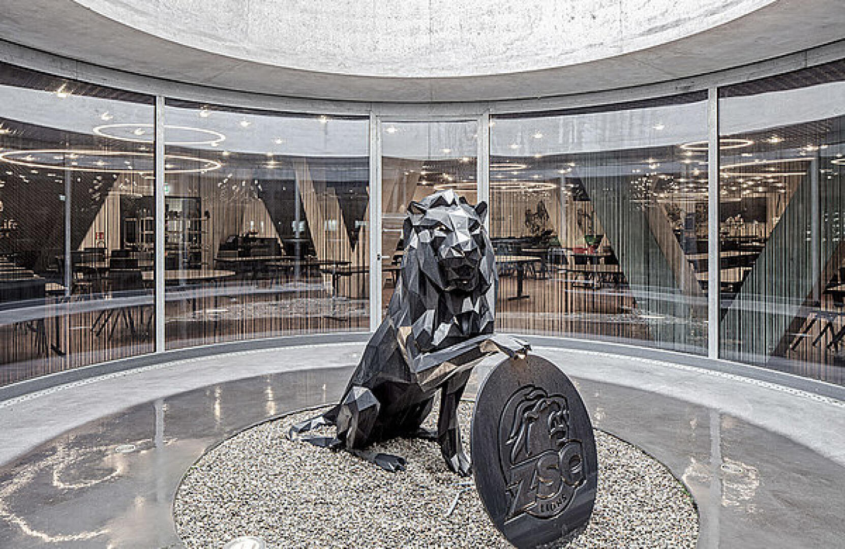 Une statue de lion noir, moderne et géométrique, repose sur du gravier dans une pièce circulaire aux parois vitrées. Le lion tient une grande plaque en forme de pièce de monnaie ornée d'une tête de lion et de l'inscription « ZSO ». Le mobilier de bureau est visible à travers les parois vitrées.