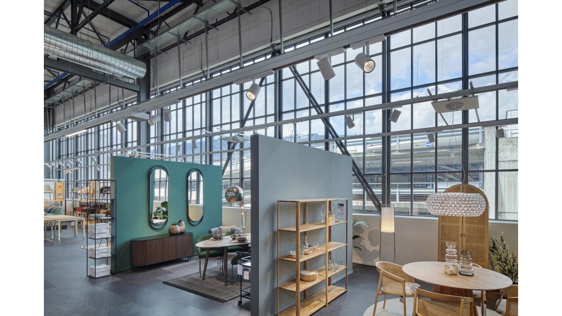 Salle d'exposition moderne et spacieuse avec de grandes fenêtres de style industriel, présentant des meubles bien rangés, notamment une table à manger, des étagères, des miroirs et des articles de décoration, dans un cadre bien éclairé et aéré.