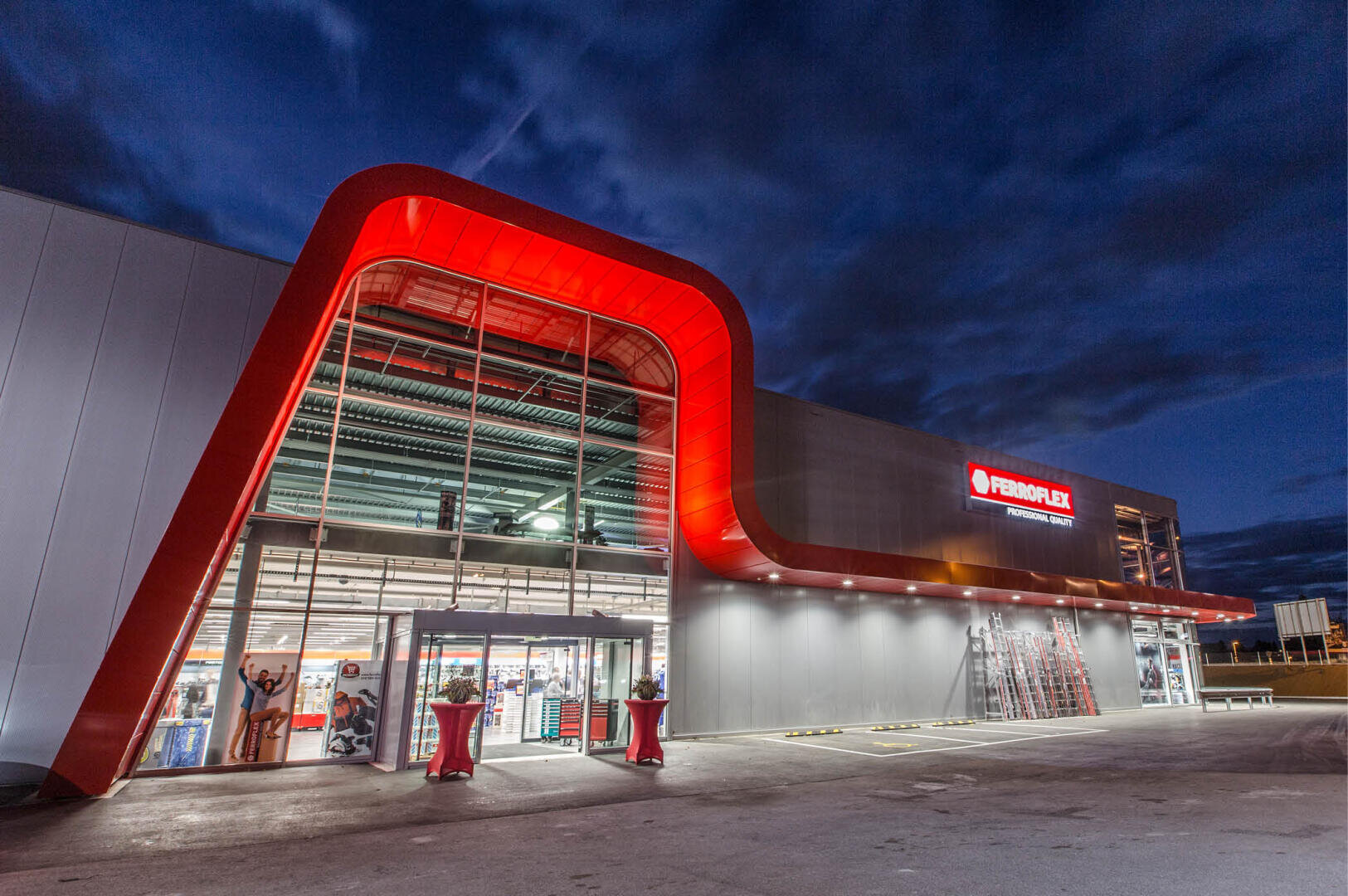 Un grand bâtiment commercial moderne, avec une caractéristique architecturale rouge frappante et une entrée en verre, est illuminé au crépuscule. Le panneau au-dessus de l'entrée indique FERROFLEX. Le ciel est sombre et nuageux.