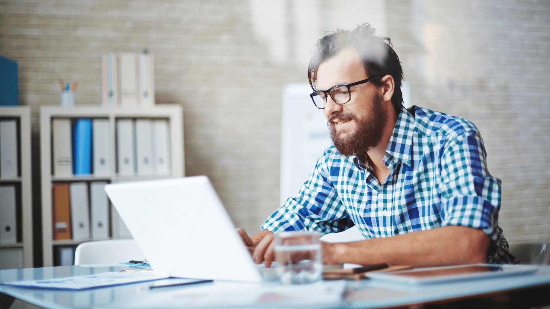 Ein bärtiger Mann mit Brille und blau-kariertem Hemd arbeitet an einem Laptop an einem Schreibtisch in einem modernen Büro, mit Regalen voller Aktenordner im Hintergrund und einem Glas Wasser auf dem Schreibtisch.