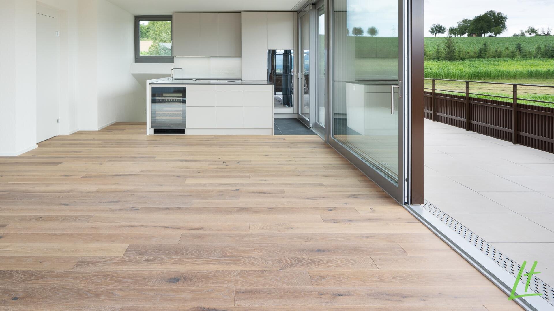Moderne, unmöblierte Küche mit hellem Holzfußboden, weißen Schränken und großen Glasschiebetüren, die zu einer geräumigen Außenterrasse mit Blick auf eine grüne Landschaft führen.