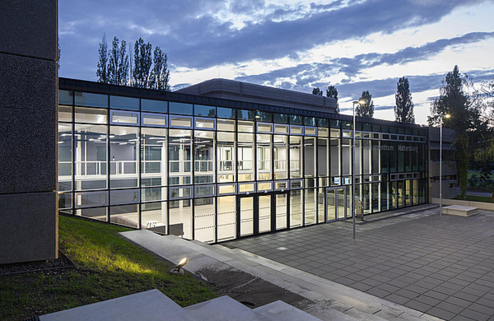 Modernes Glasgebäude mit großen, von innen beleuchteten Fenstern in der Abenddämmerung, umgeben von einem gepflasterten Innenhof und Bäumen im Hintergrund unter einem bewölkten Himmel.